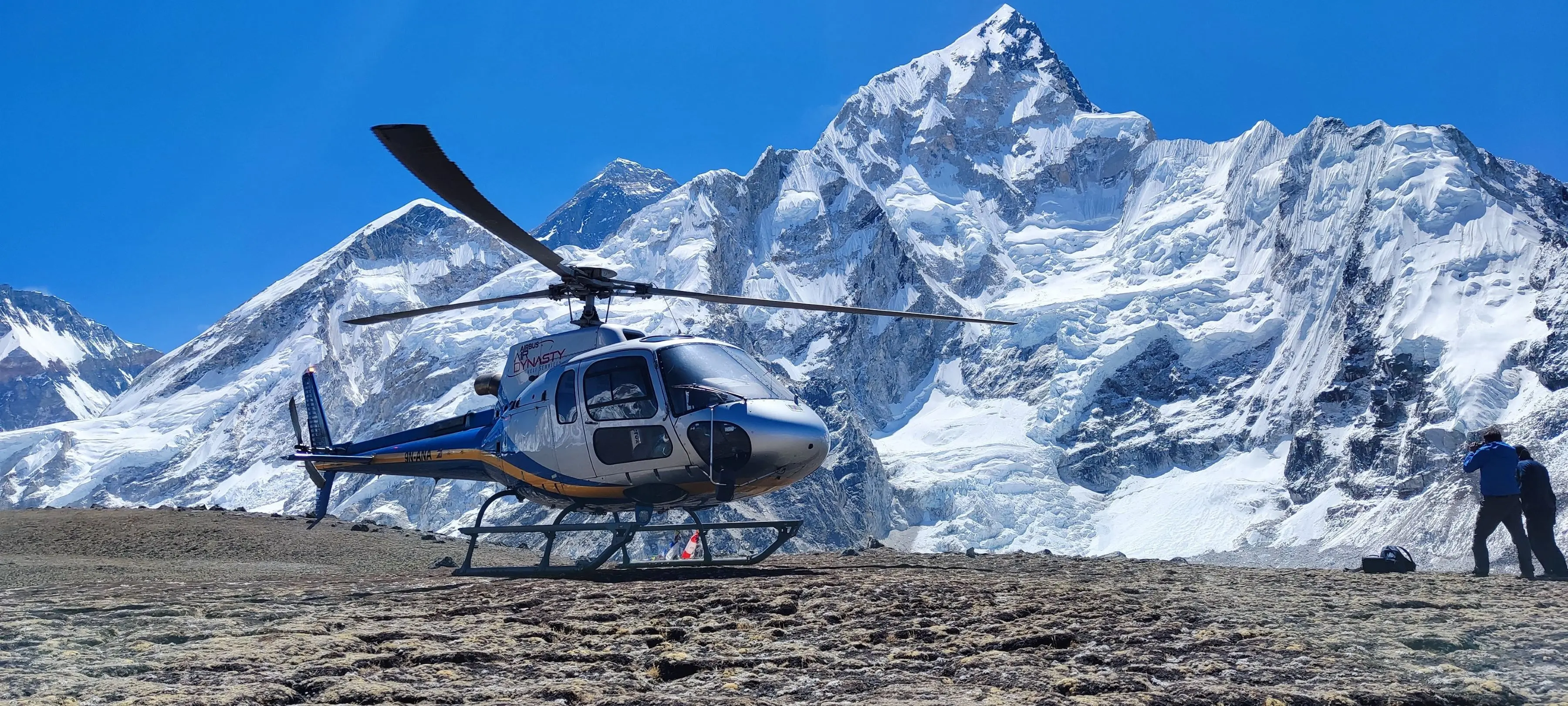Dynasty helicopter with tail number 9N-ALP, parked on a rocky high-altitude landing pad, with a spectacular detailed panorama of massive, snow-covered Himalayan peaks