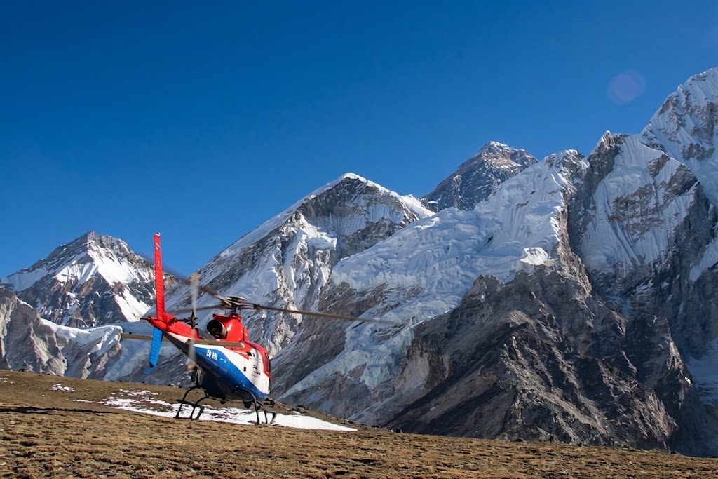 A red, white, and blue helicopter landed on a patch of snow and rocky ground. Behind it, the monumental, snow-capped peaks of the Everest region massif, with detailed ice and rock faces.